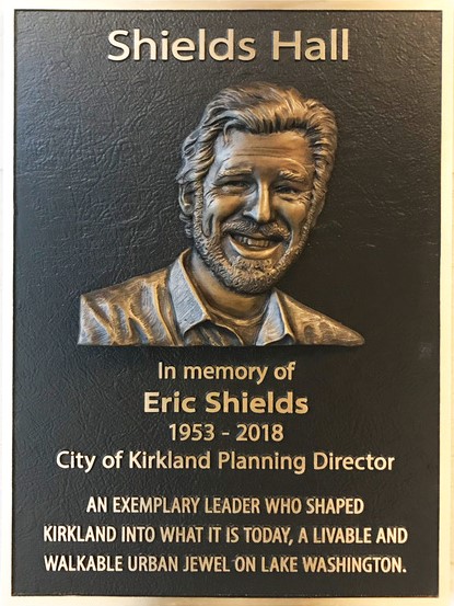 Bronze Plaque with a picture of a man with medium length swept back hair, a big smile, and a beard. The plaque reads: "Shields Hall In memory of Eric Shields 1953-2018 City of Kirkland Planning Director - An exemplary leader who shaped Kirkland into what it is today, a livable and walkable urban jewel on Lake Washington"
